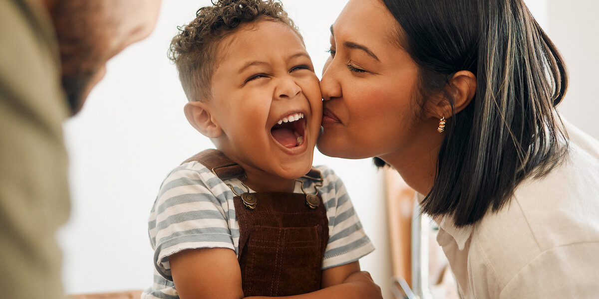 Happy boy getting a kiss by caring mother, bonding and laughing during family time at home. Young parents sharing a sweet moment of parenthood with their playful child, relaxing and carefree together Happy boy getting a kiss by caring mother, bonding and laughing during family time at home. Young parents sharing a sweet moment of parenthood with their playful child, relaxing and carefree together