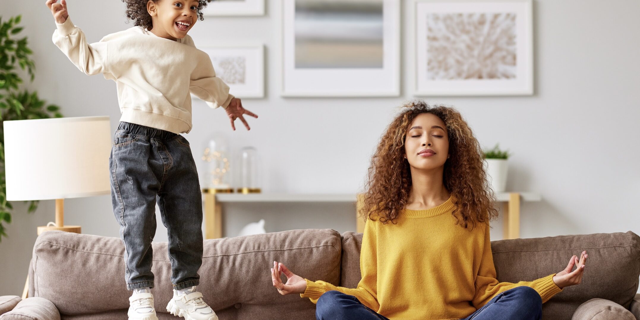 Young african woman mother in lotus pose meditating while child playing on background in living room Young african woman mother in lotus pose meditating while child playing on background in living room